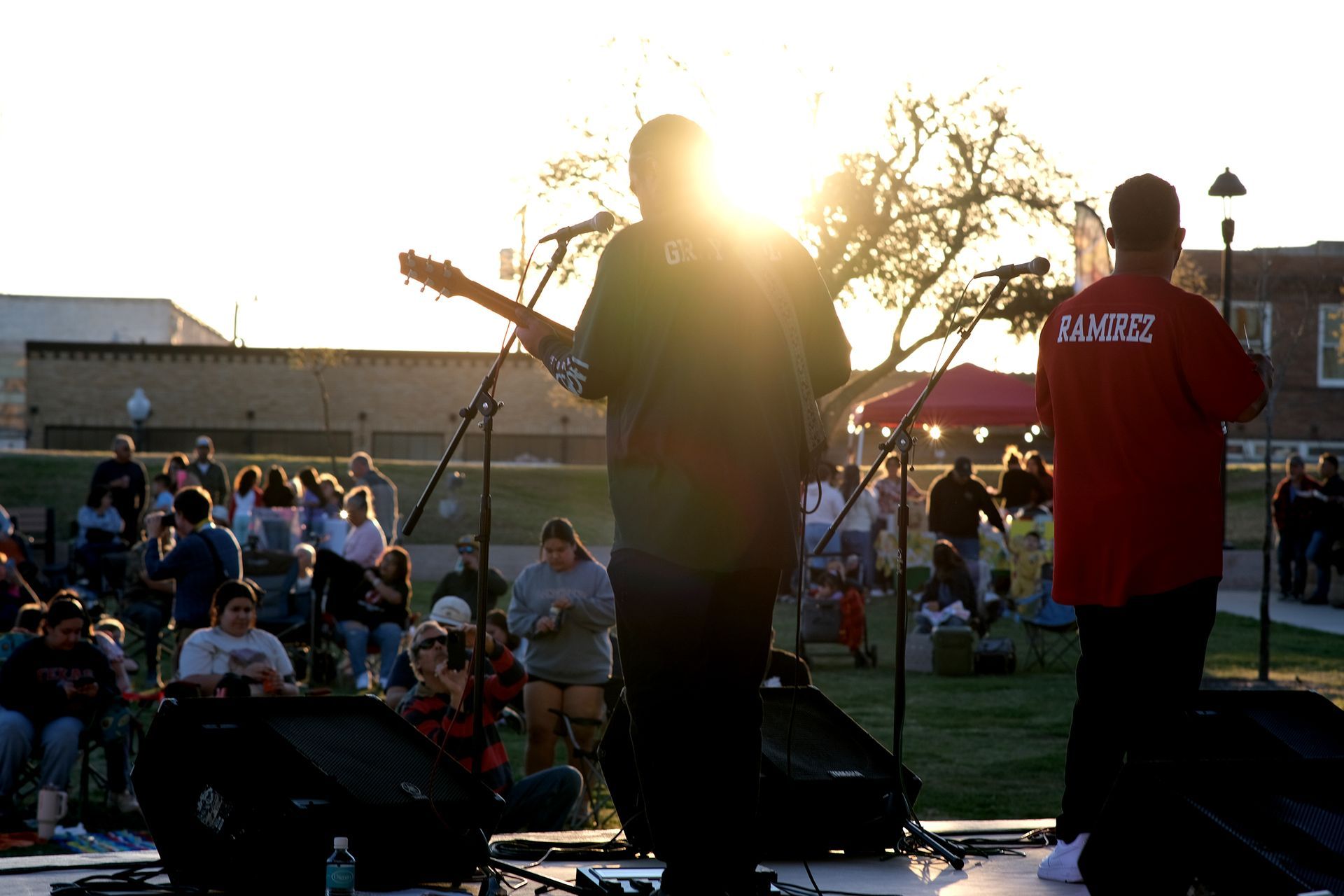 Band performs on outdoor stage, backlit by sunset. Audience watches on a grassy area.