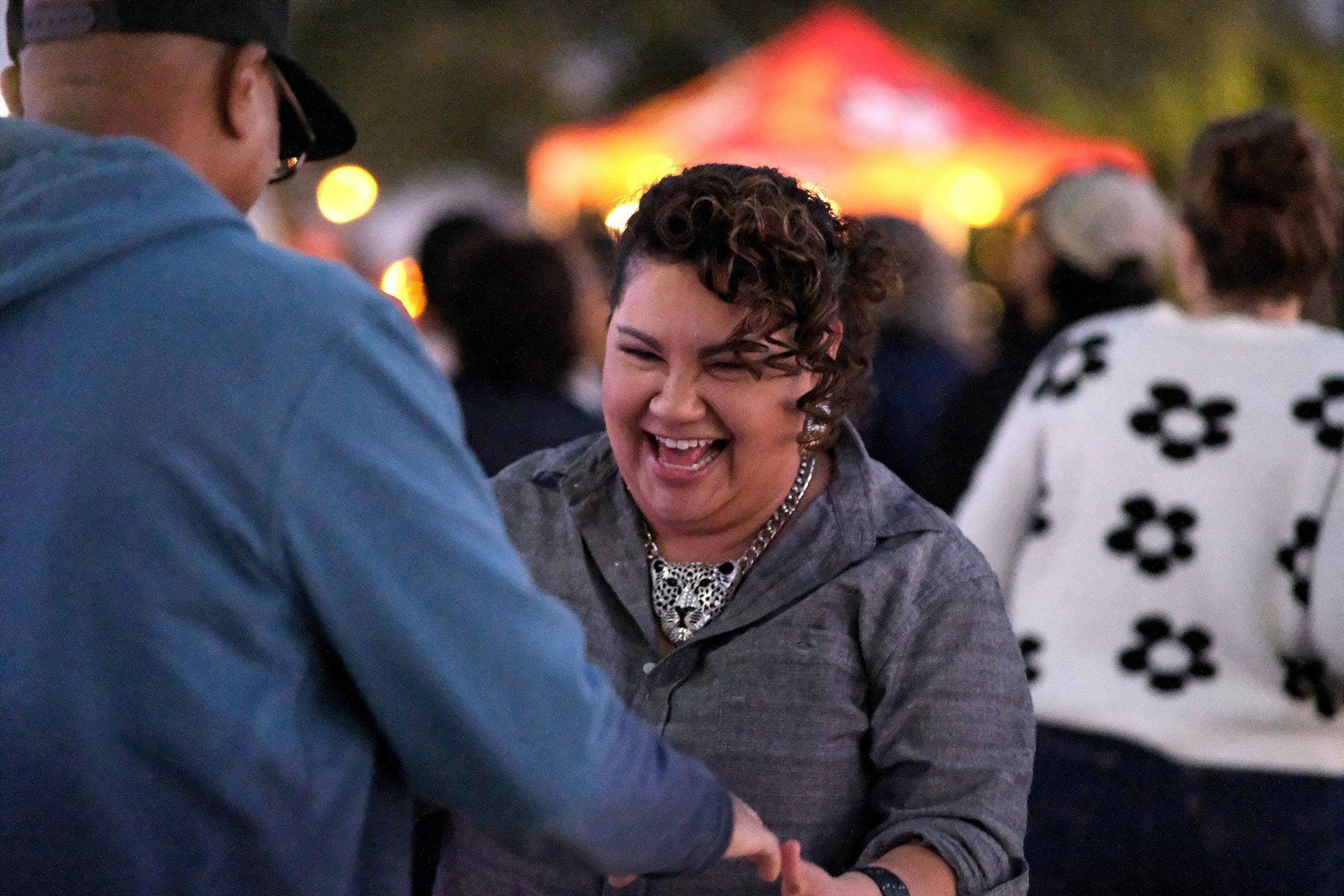 Woman laughing with open mouth, shaking hands with a person wearing a blue hoodie at an outdoor gathering.