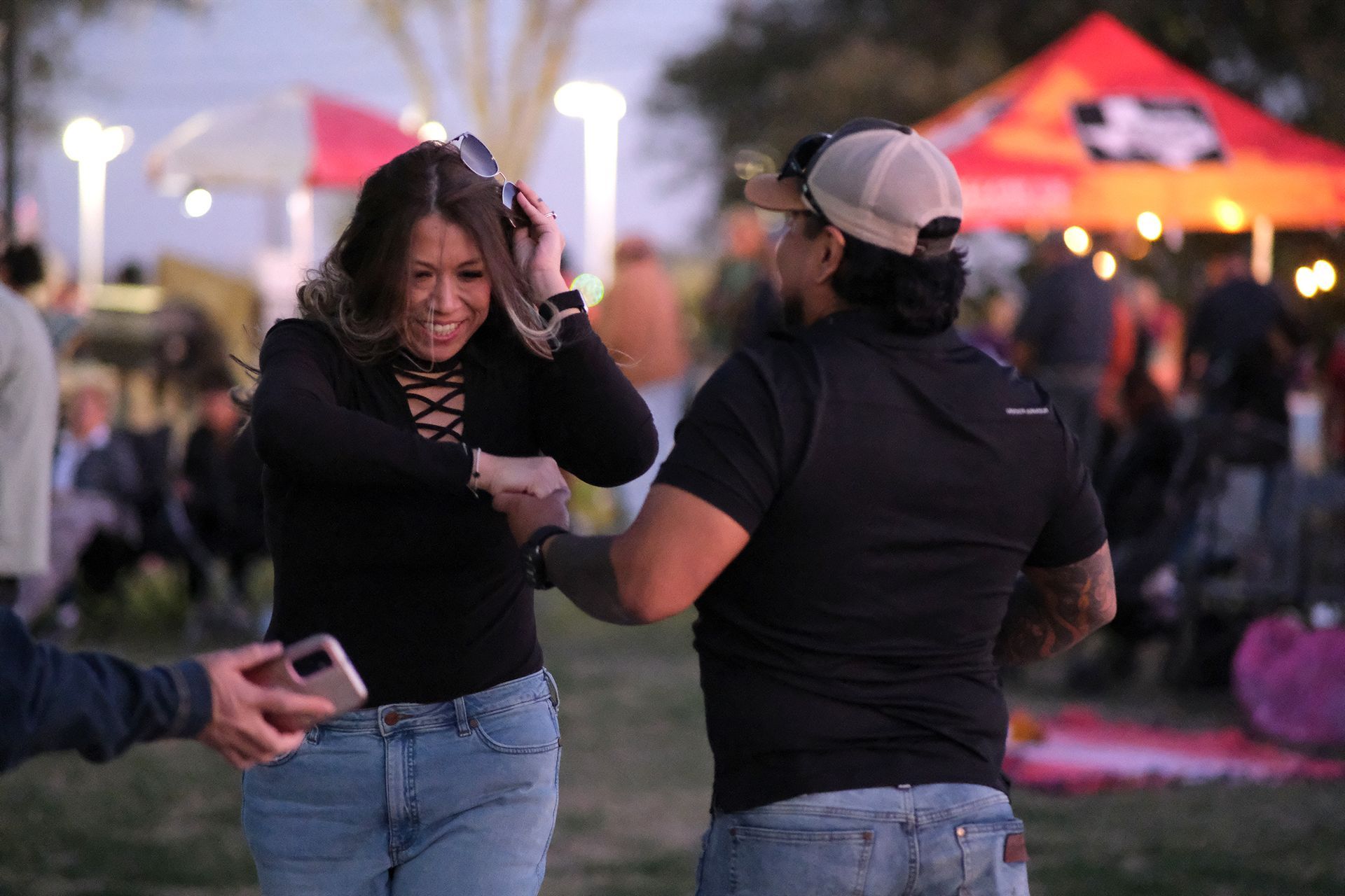 Woman dancing outdoors with a man, people in background, soft lighting.