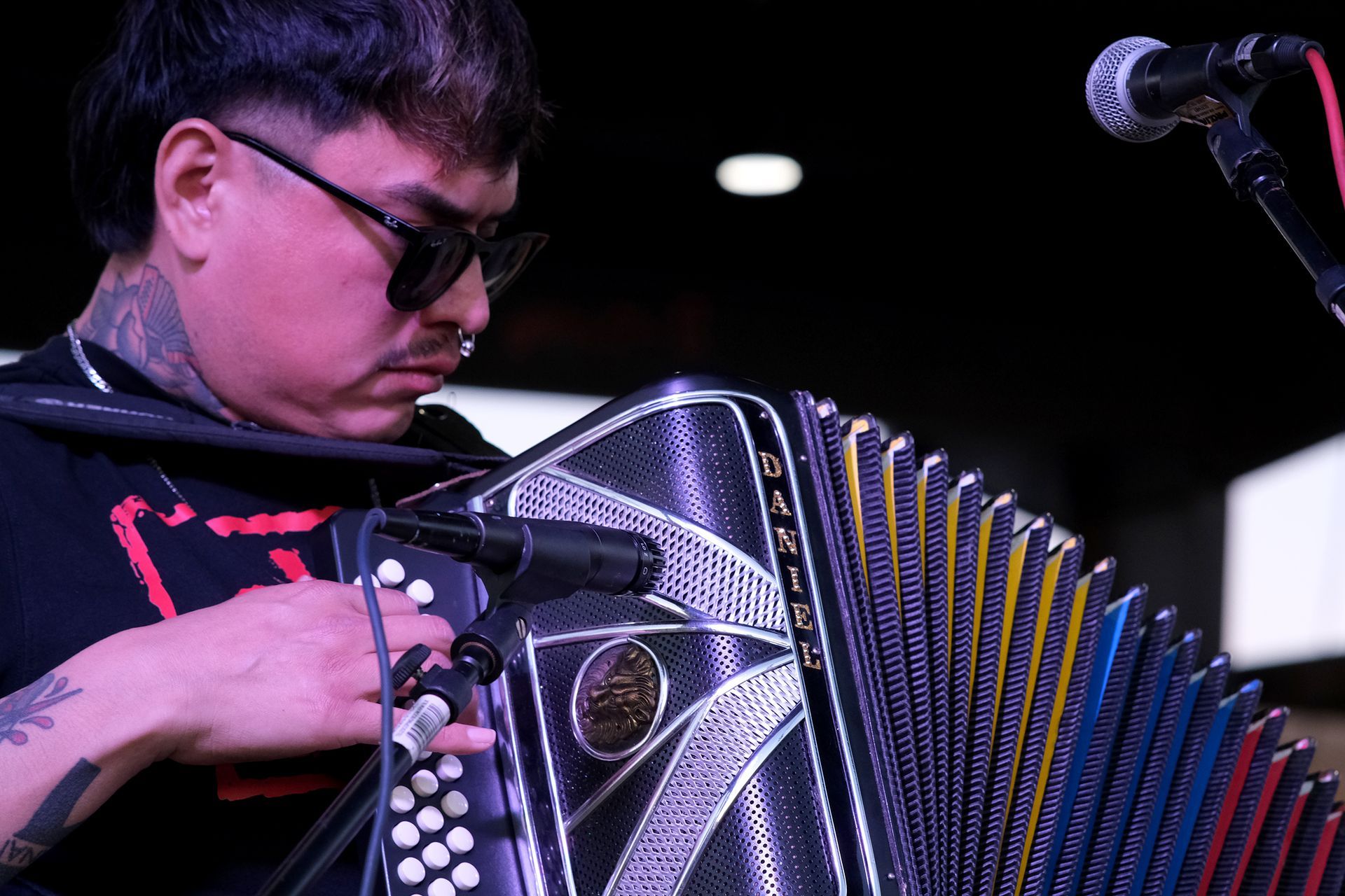 Man playing an accordion into a microphone. Dark glasses, tattoos, indoors.