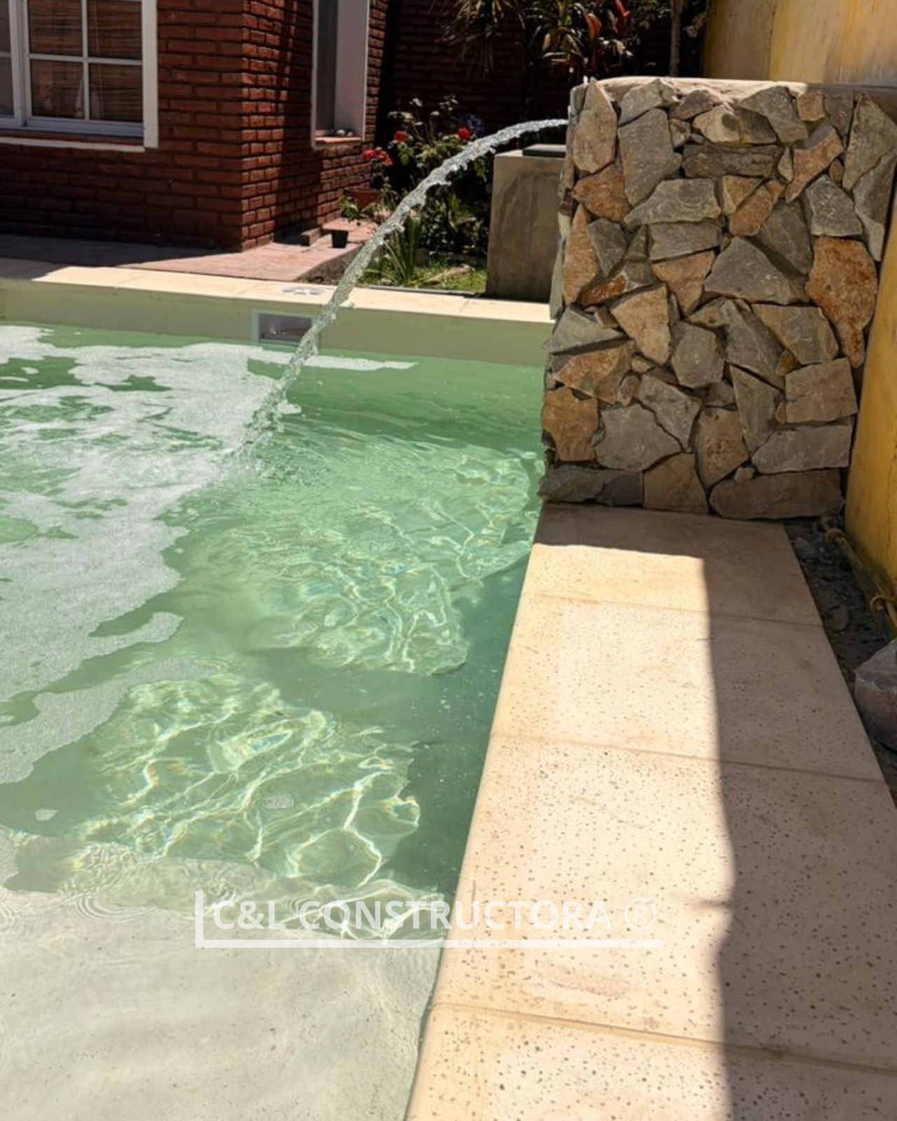 Piscina con fuente de piedra, hormigón beige y un edificio de ladrillo al fondo.