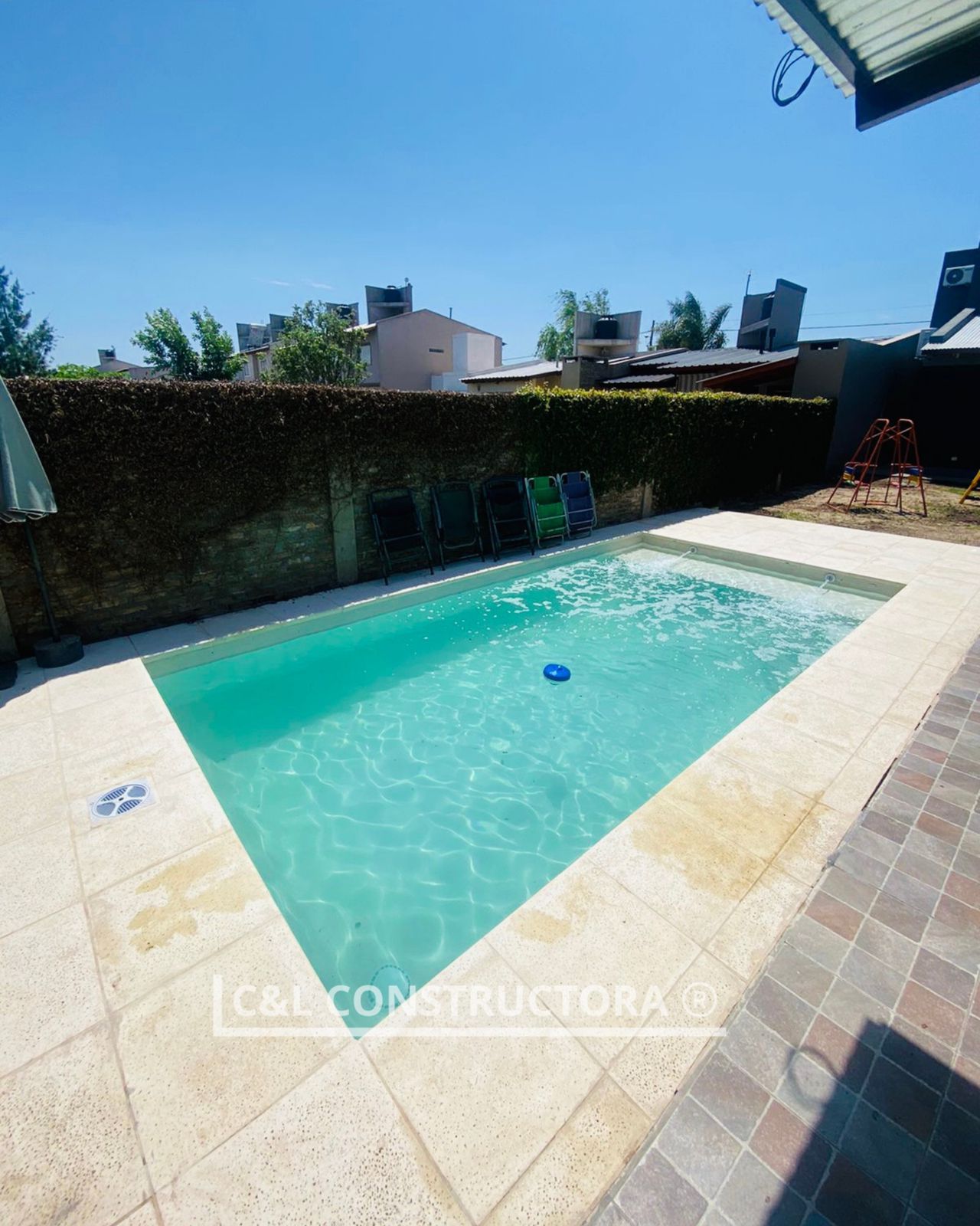 Una piscina rectangular rodeada de baldosas de piedra. Un seto verde al fondo bajo un cielo azul.