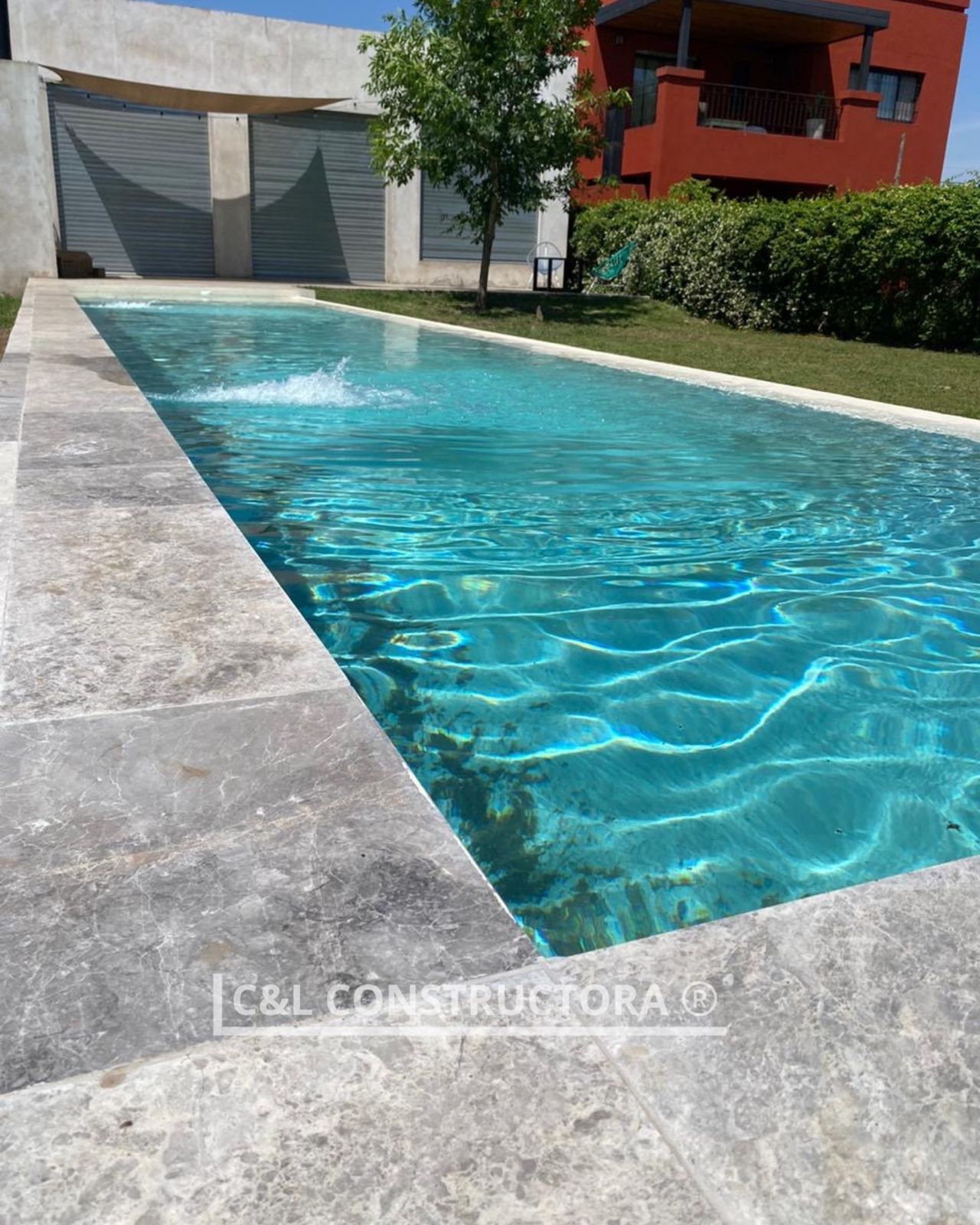 Piscina rectangular de agua turquesa, borde de piedra y fuente. Casas y césped al fondo.