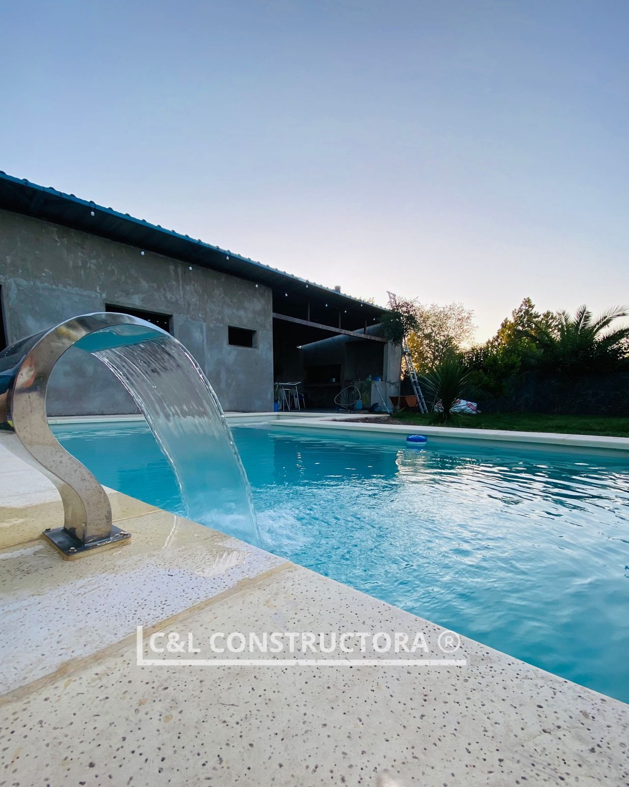 Piscina con cascada y edificio al fondo. Agua azul cristalina y un marco de piedra clara.