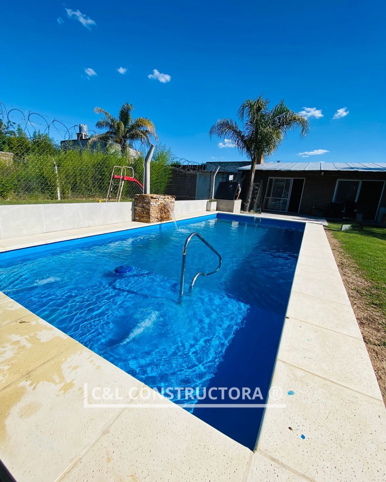 Piscina rectangular de color azul con agua, azulejos circundantes de color claro y un cielo soleado.