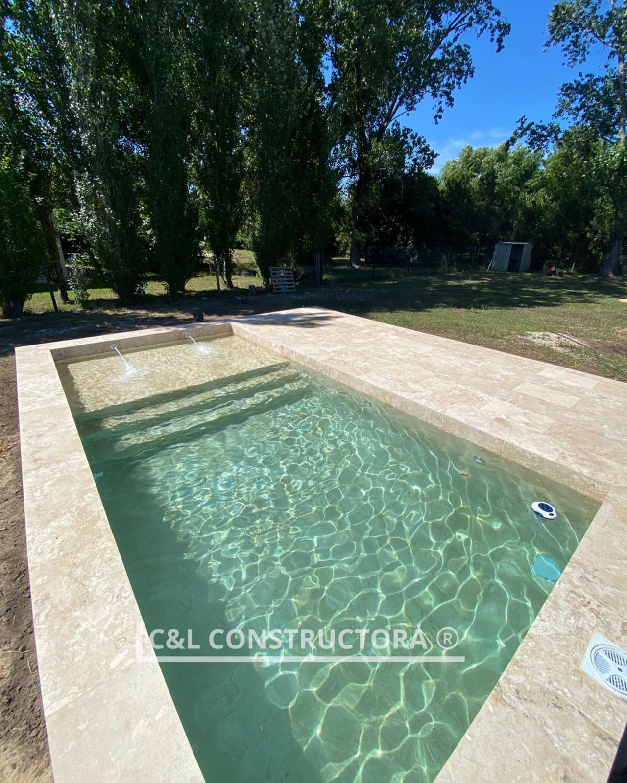 Piscina rectangular con borde de piedra clara; el agua cristalina refleja el cielo. Rodeada de césped y árboles.