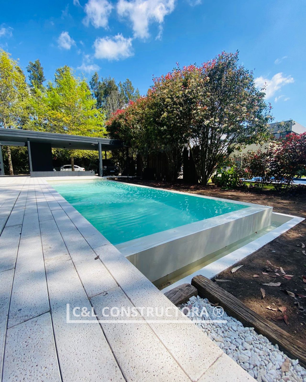 Piscina rectangular de agua turquesa, rodeada de piedra blanca y exuberante vegetación bajo un cielo azul.