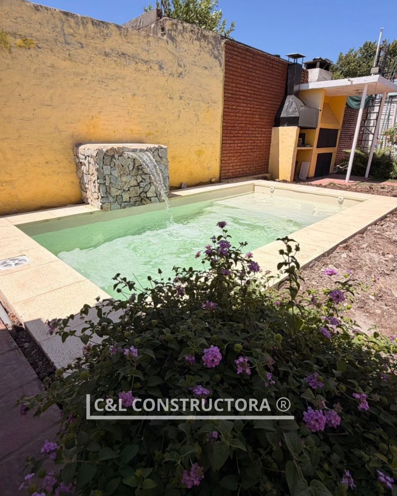 Pequeña piscina al aire libre con cascada, bordeada de piedra beige y una pared amarilla, con paisajismo.