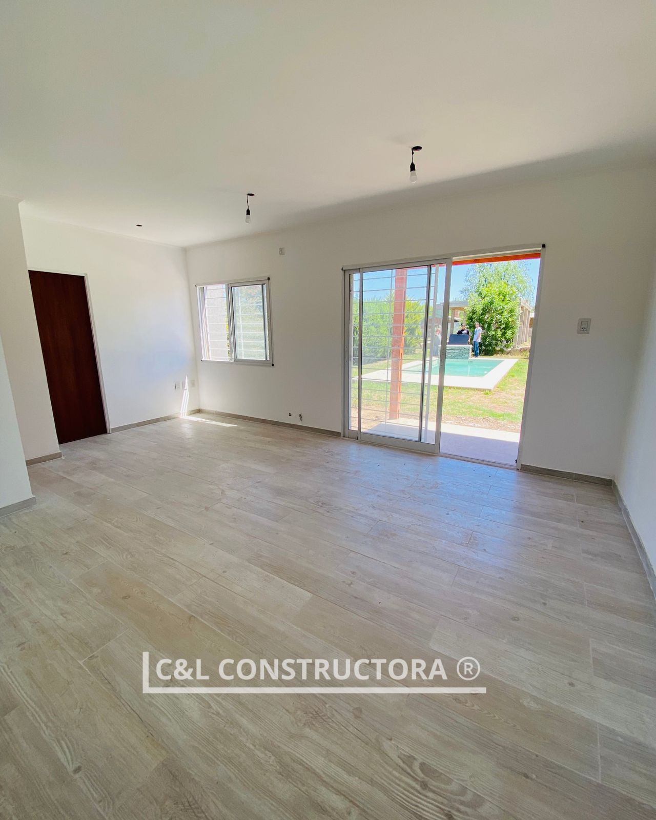 Habitación vacía con pisos de madera clara, paredes blancas y una puerta corrediza de vidrio a un patio trasero con piscina.