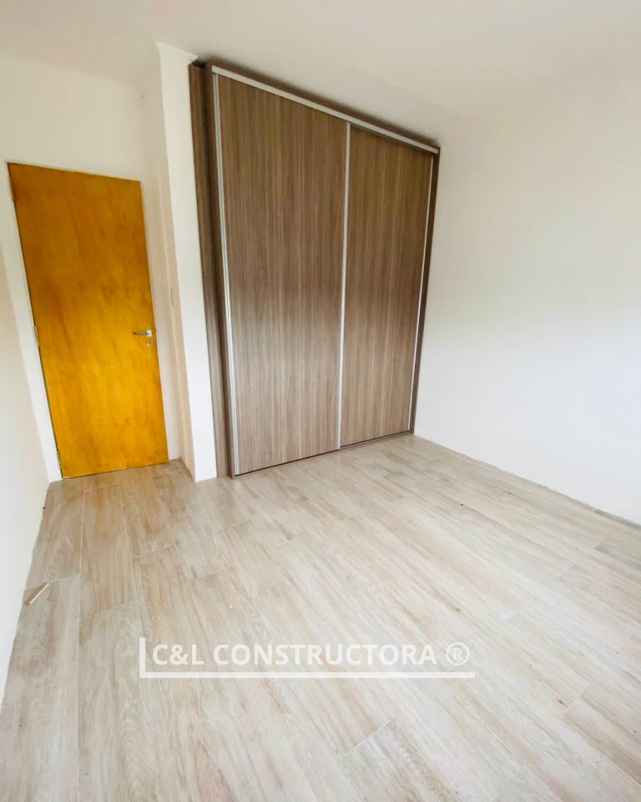 Dormitorio vacío con piso de apariencia madera, puertas de armario de madera y una puerta de color dorado.
