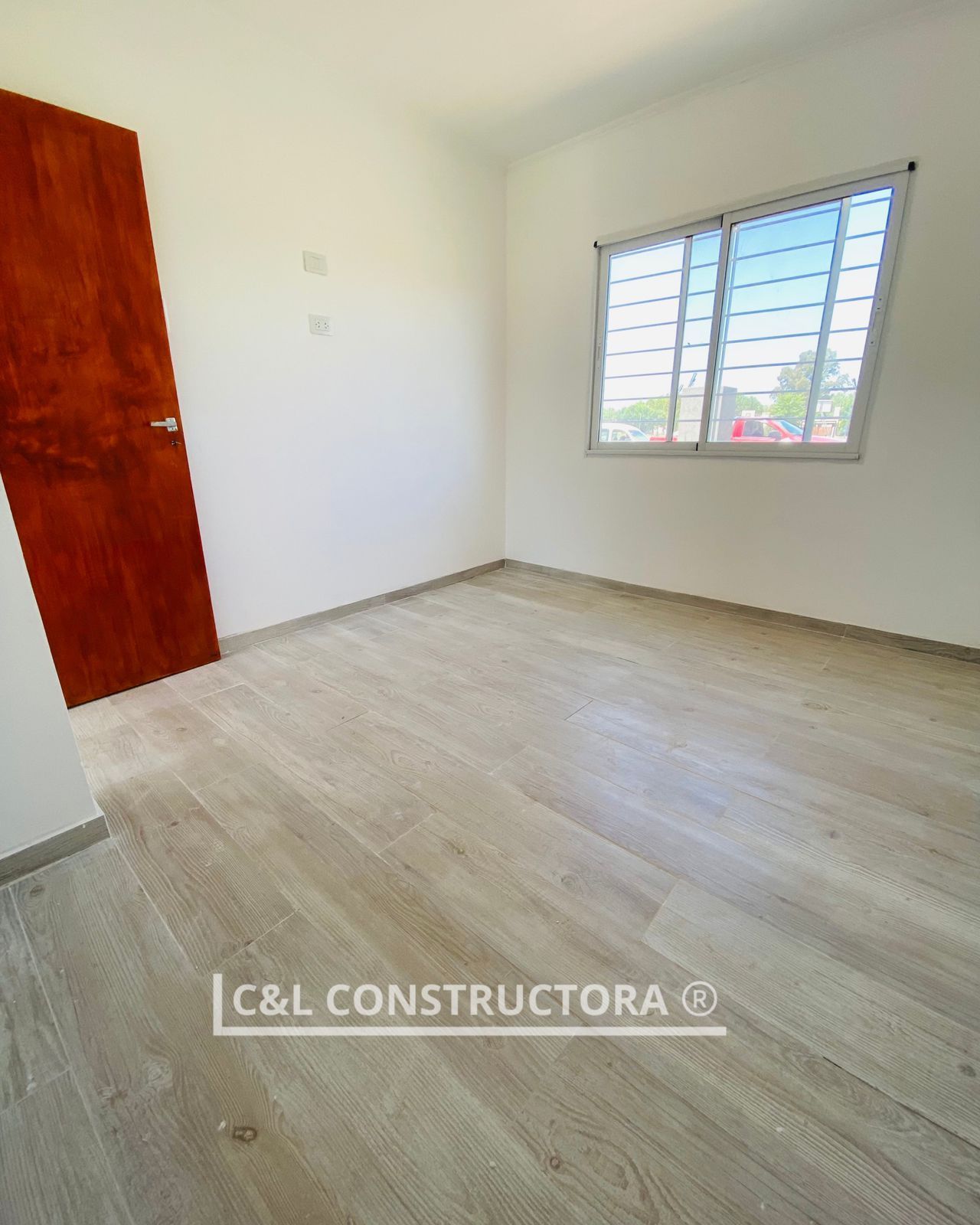 Habitación vacía con suelo de madera clara, paredes blancas, puerta y ventana oscuras.