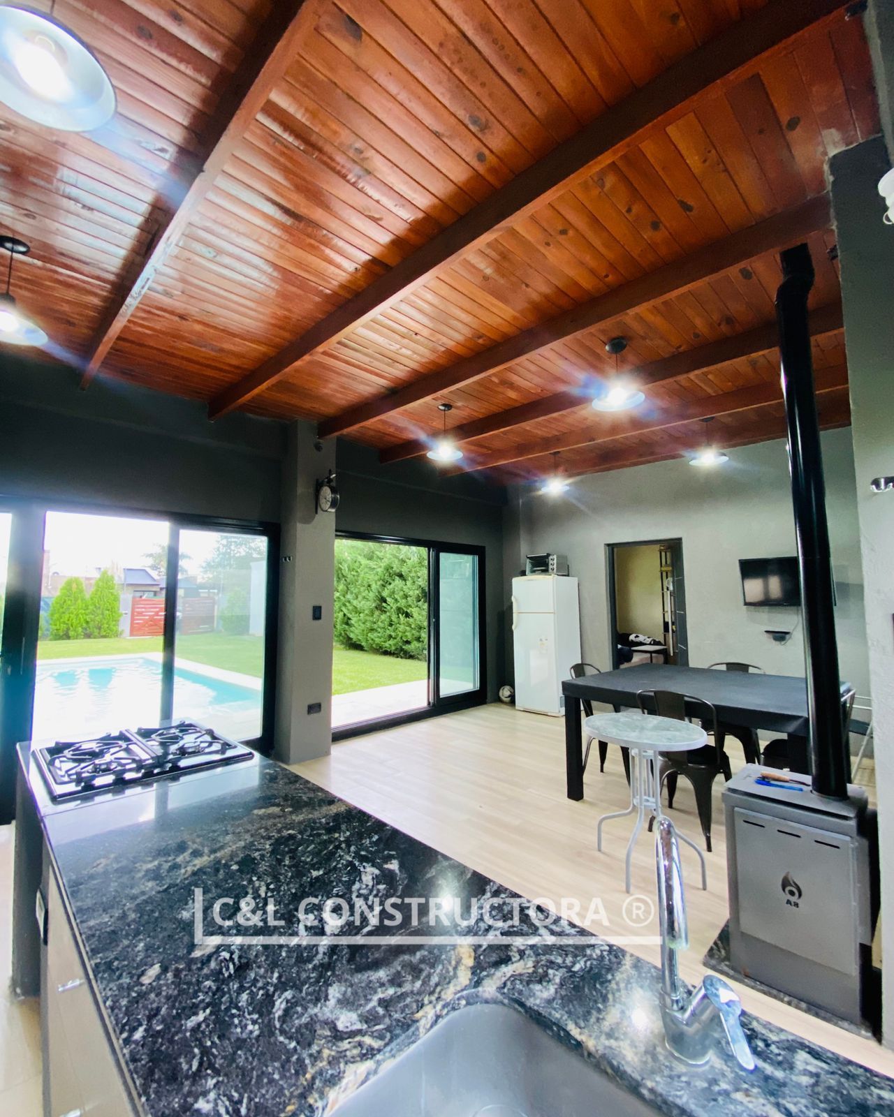 Interior de una cocina moderna con techo de madera, encimeras oscuras y vista a una piscina.