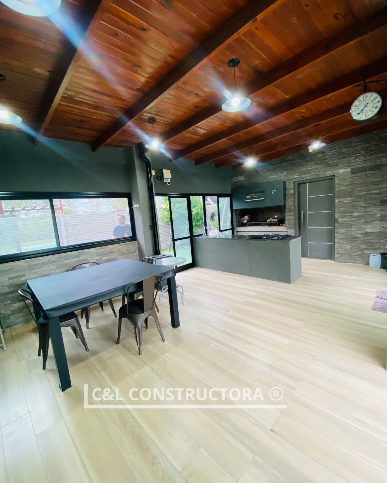 Cocina-comedor de concepto abierto con techo de madera, gabinetes grises, ventanas grandes y una mesa larga y oscura.