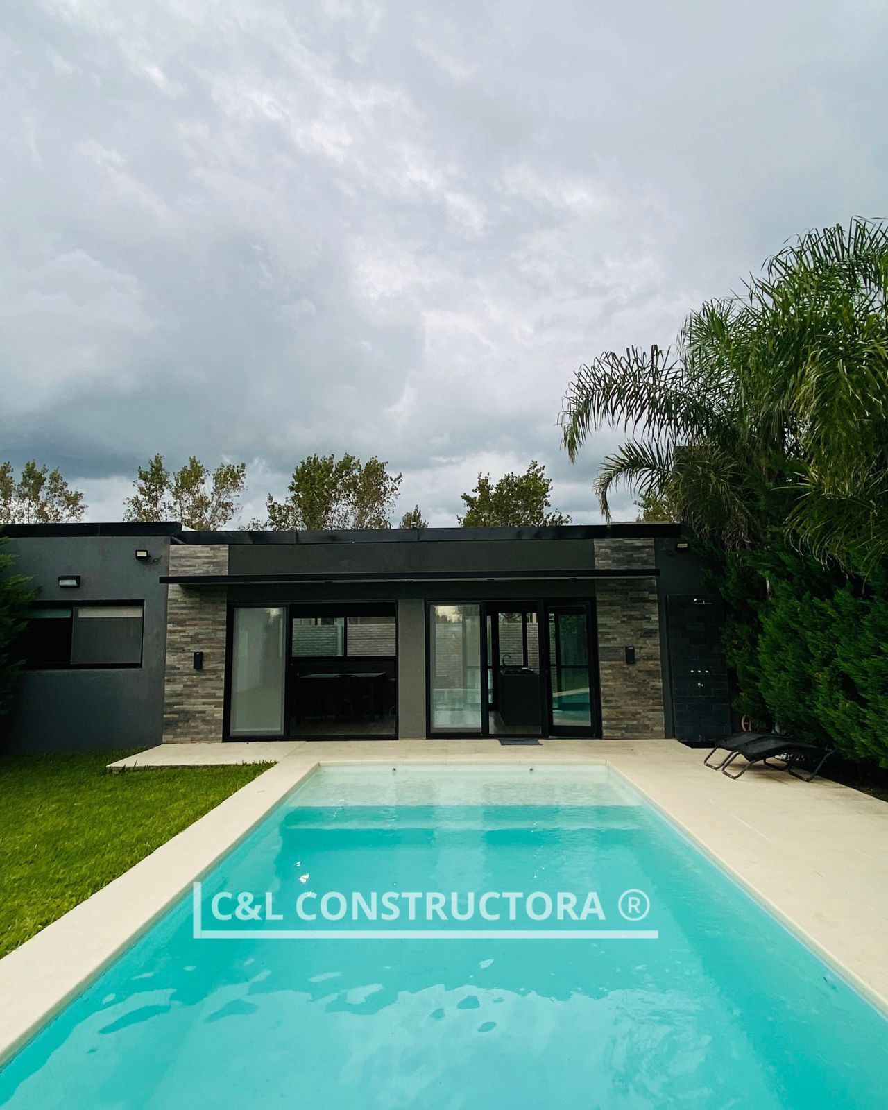 Una casa moderna con piscina. El edificio tiene paredes oscuras, fachada gris y puertas de cristal. Cielo nublado.