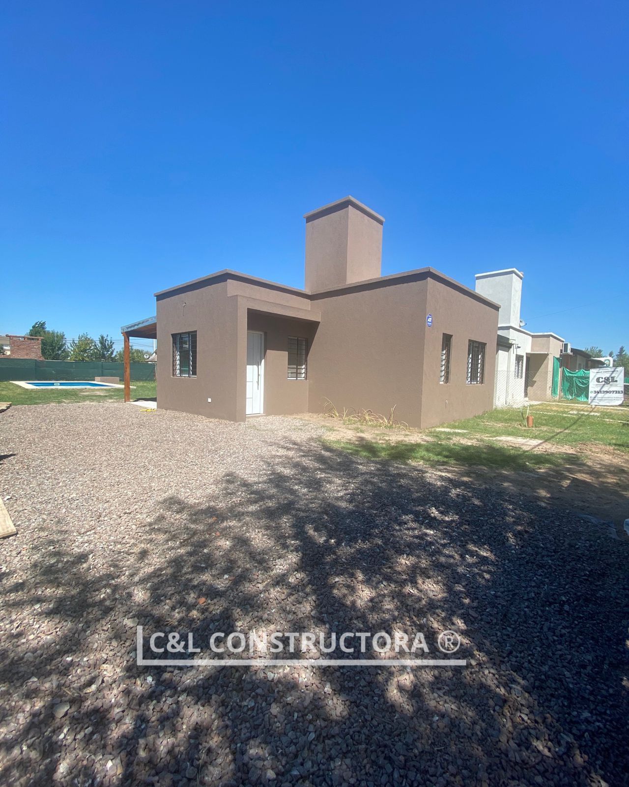 Casa marrón en construcción con chimenea y puerta blanca contra un cielo azul. Patio de grava.