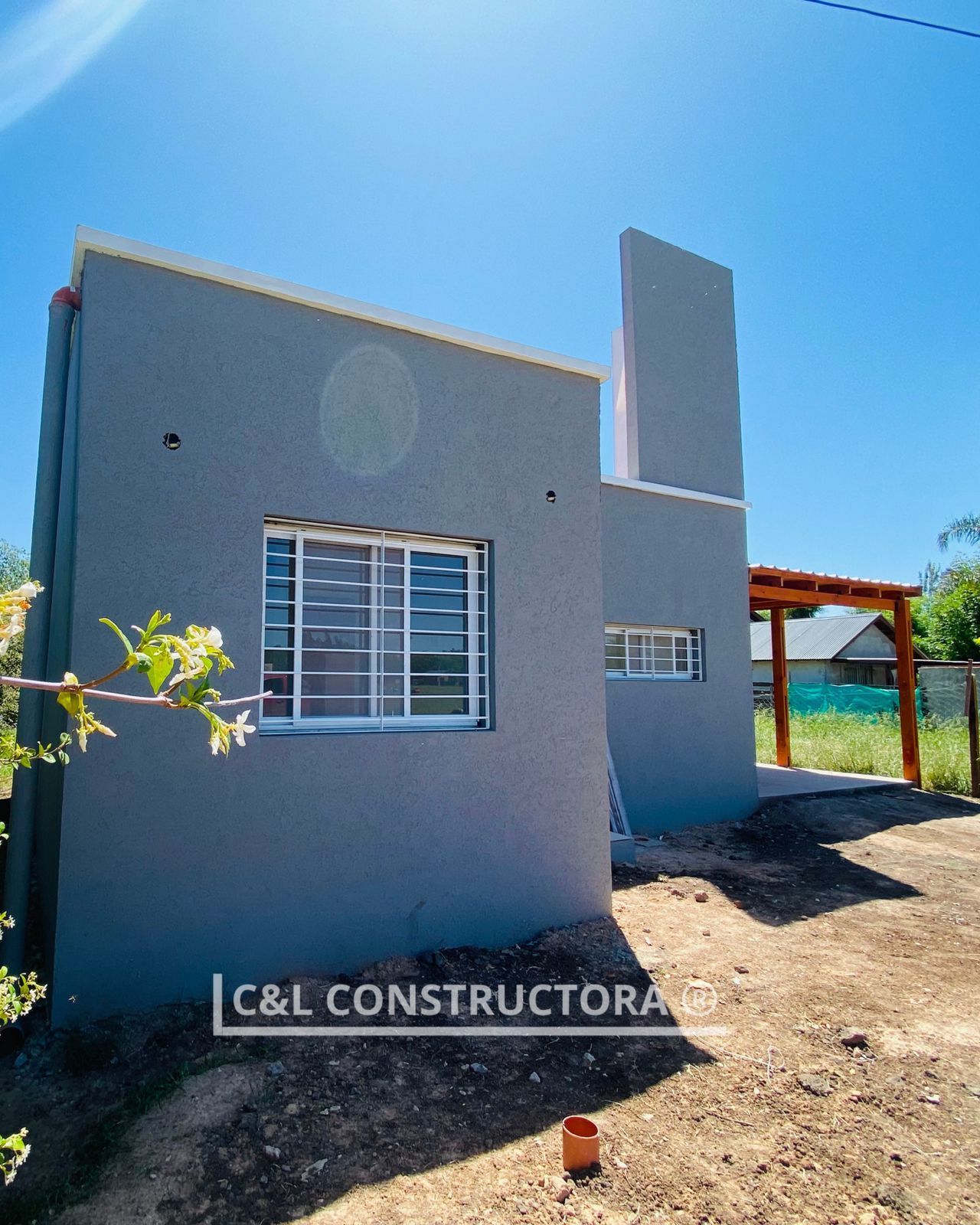Casa de estuco gris con ventanas de marco blanco, pérgola y cielo azul.