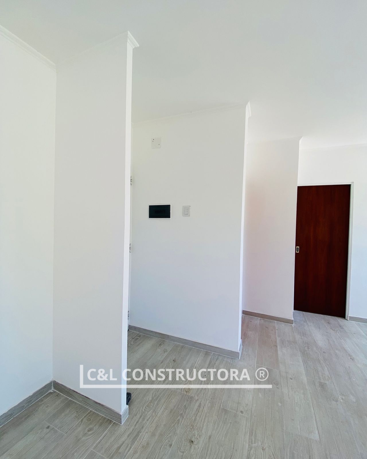 Vista interior de una habitación de nueva construcción con paredes blancas, puerta de madera y piso de madera clara.