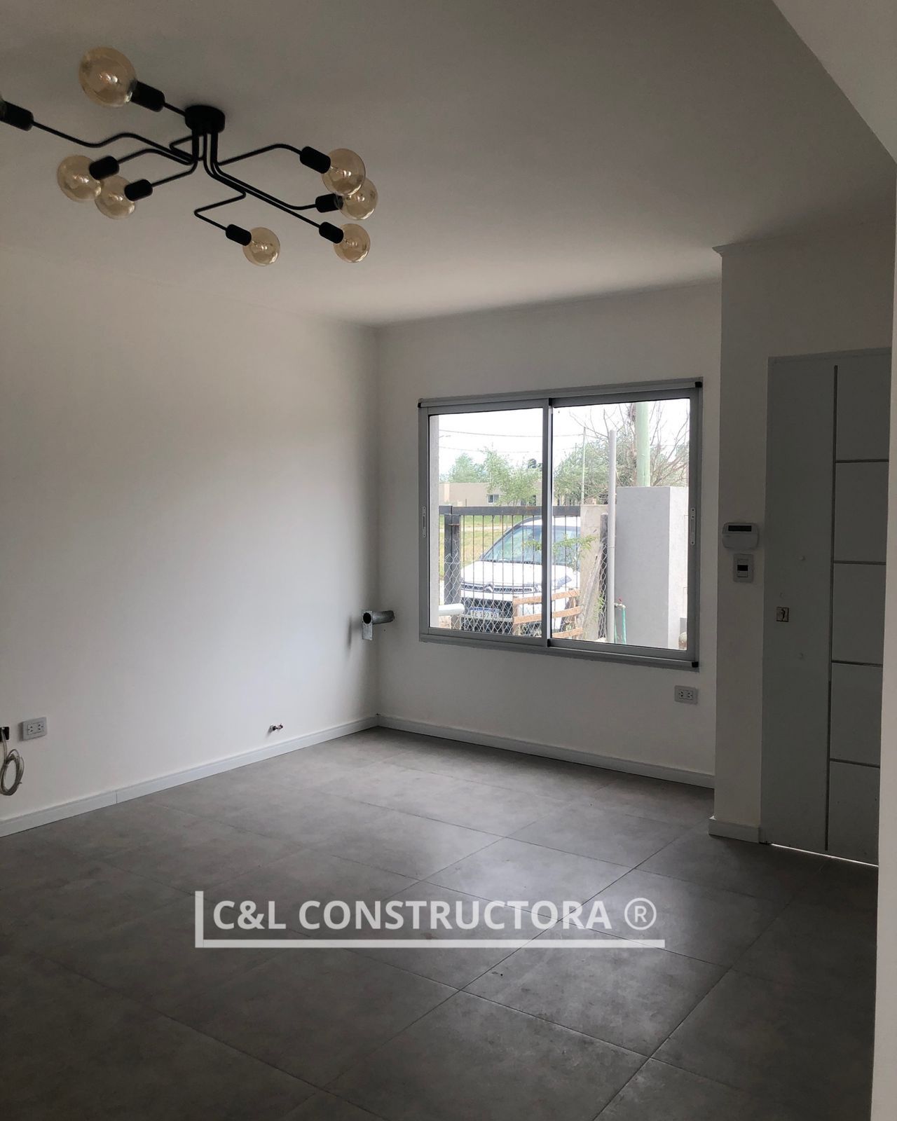 Habitación vacía con suelo de cemento, paredes blancas, ventana y una lámpara de araña moderna.