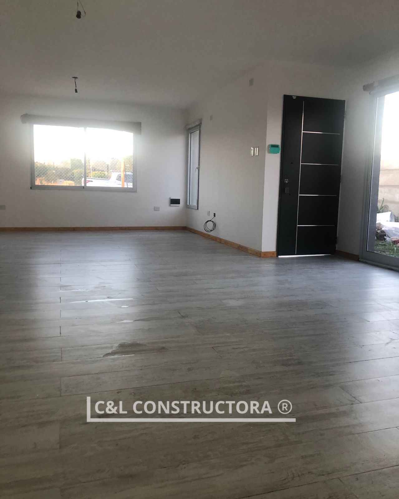 Interior vacío con suelo de imitación madera, ventanas y puerta negra; la luz entra a raudales.