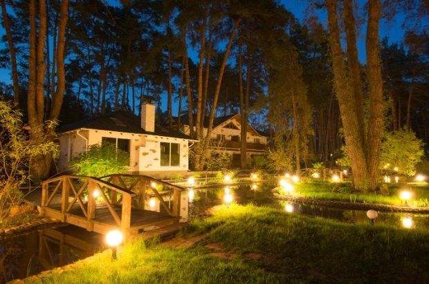 A house in the middle of a forest with a bridge over a pond at night.