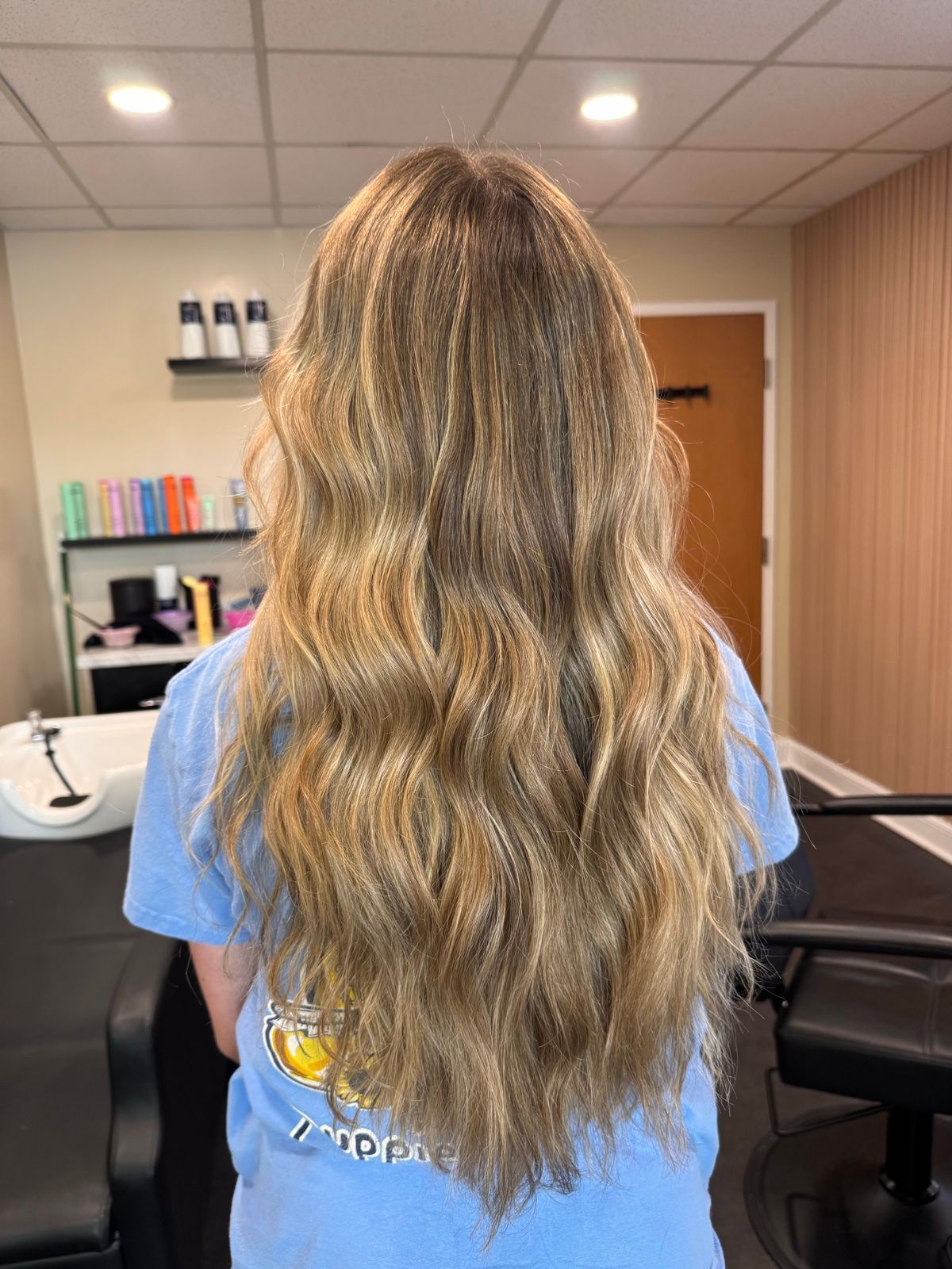 Long wavy blonde hair from behind in a salon chair, with mirrors, shelves, and warm lighting in the background