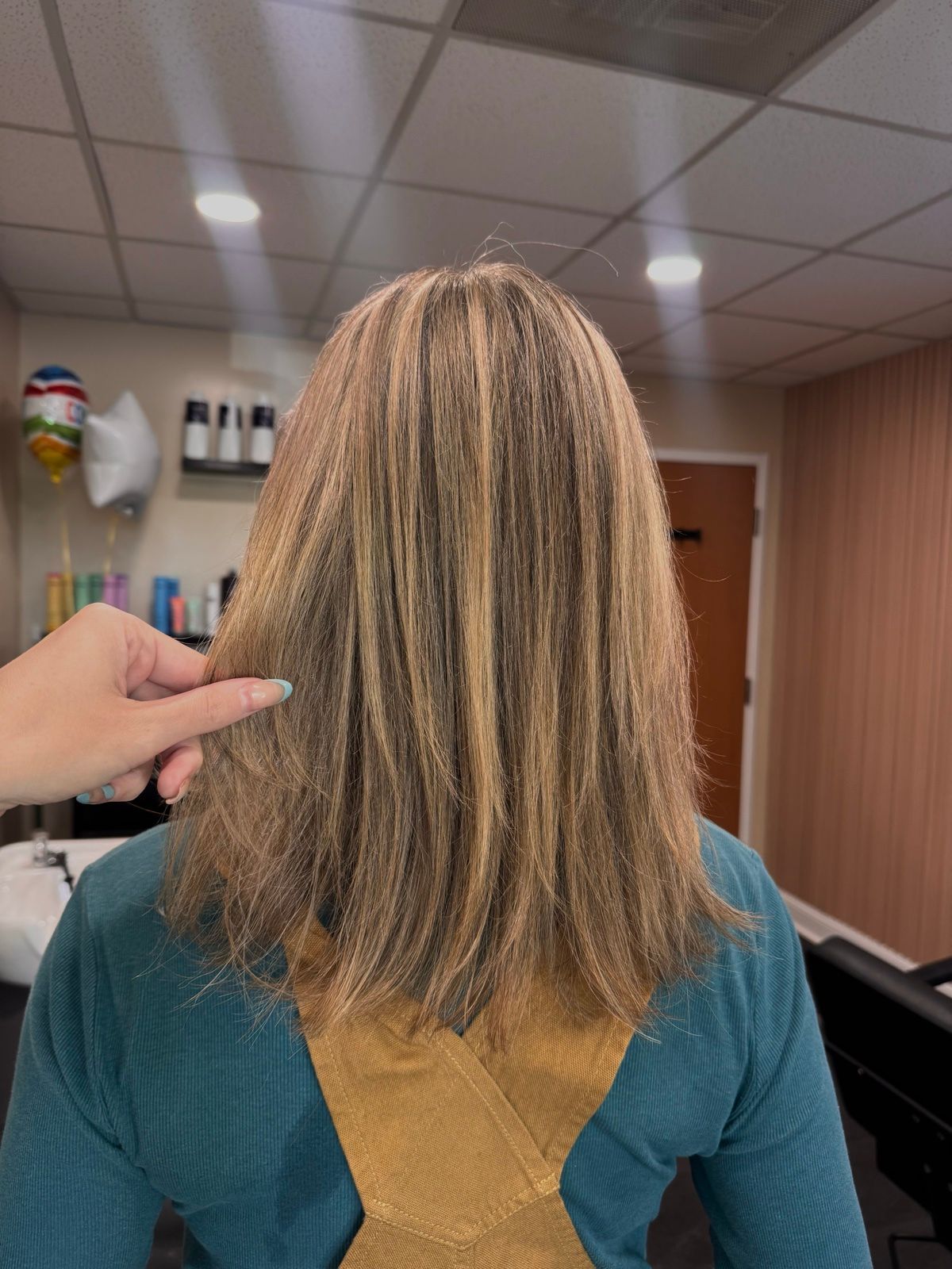 Person with highlighted hair in salon chair, back view, with a hand pointing at the hair.