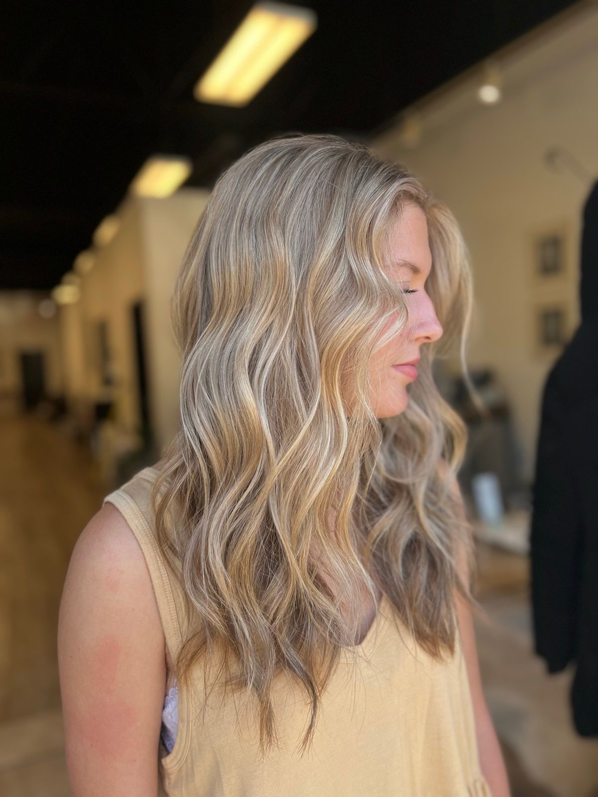 Woman with long wavy blonde hair in a yellow sleeveless top, standing indoors in a hallway.