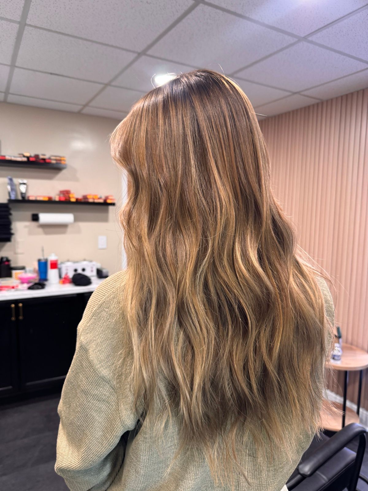 Person with long wavy hair standing in a salon, seen from behind, with shelves and products in the background