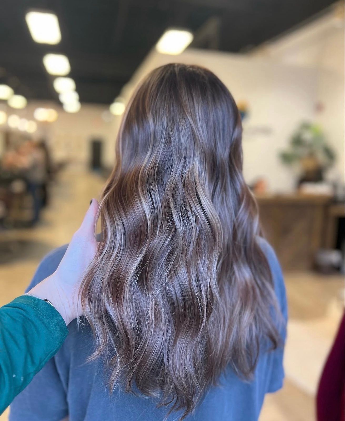 Person with long wavy brown hair seen from behind in a salon, with a hand gently touching their shoulder.