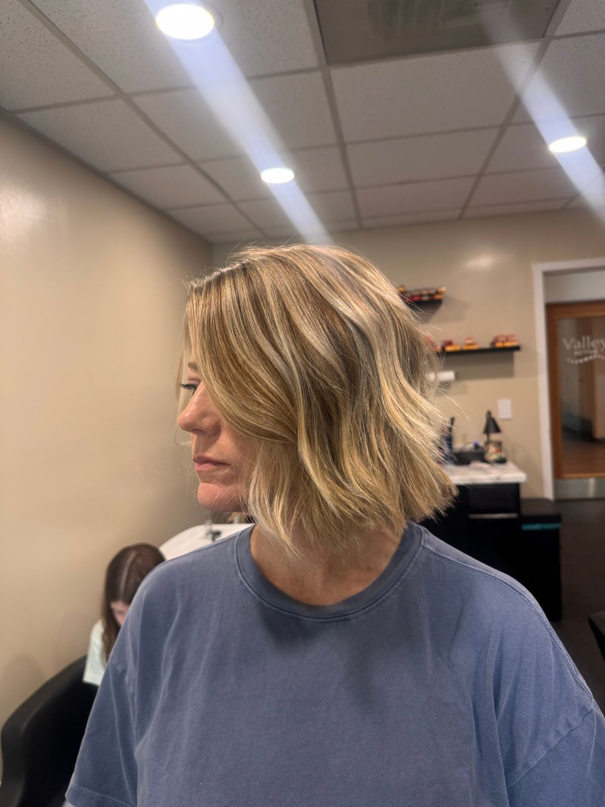 Woman in a salon, shown in profile with shoulder-length blonde hair and a blue shirt.