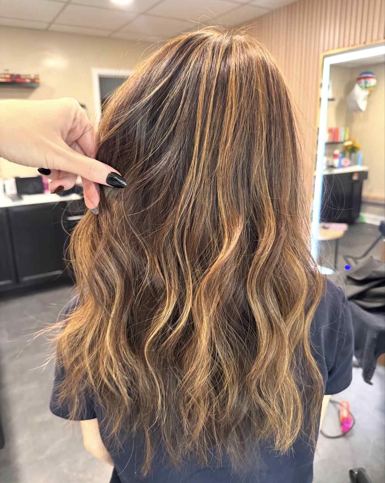 Long wavy brown hair with blonde highlights being sectioned by a hand in a salon chair
