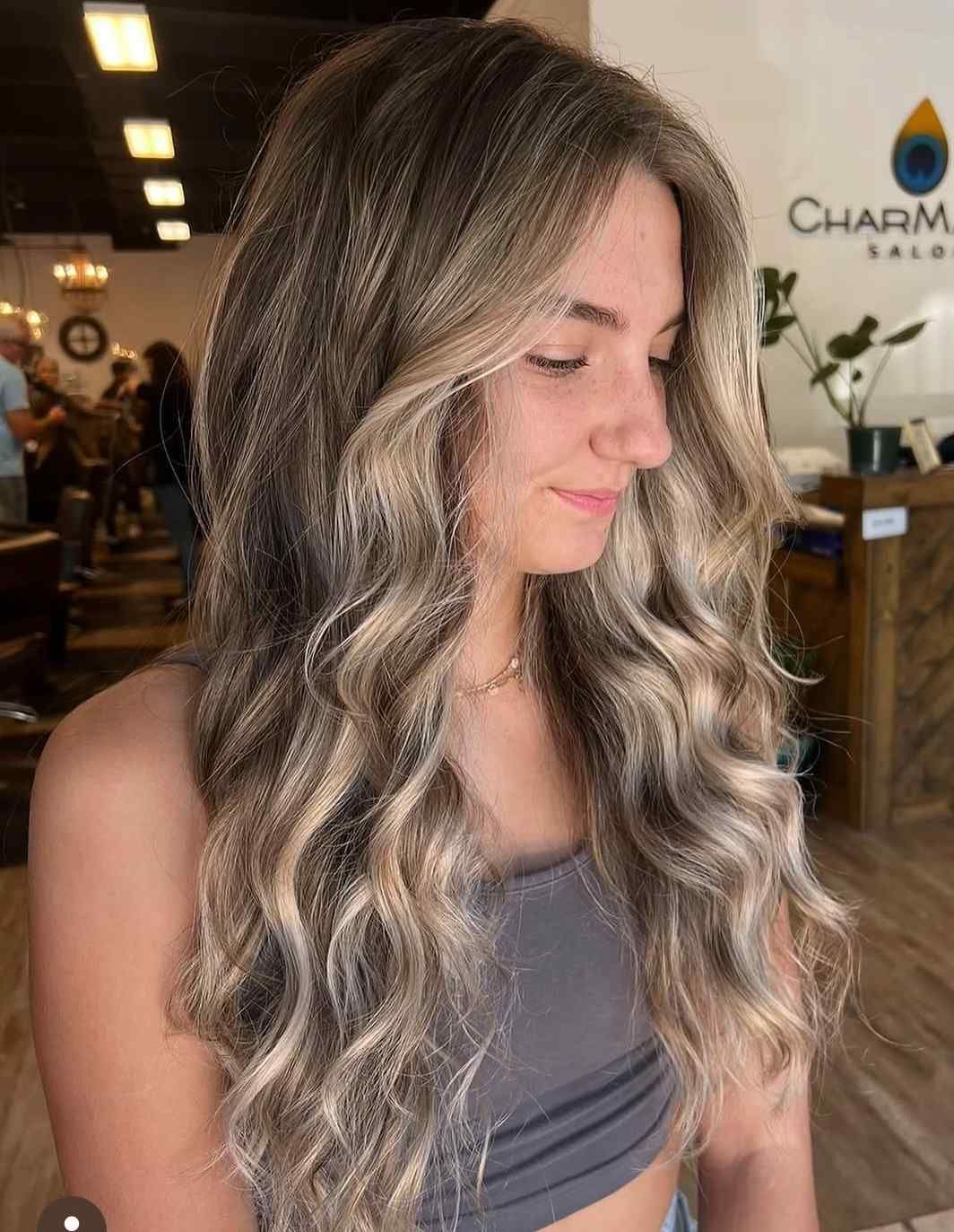 Woman with long wavy blonde hair in a salon, looking down in a sleeveless gray top.