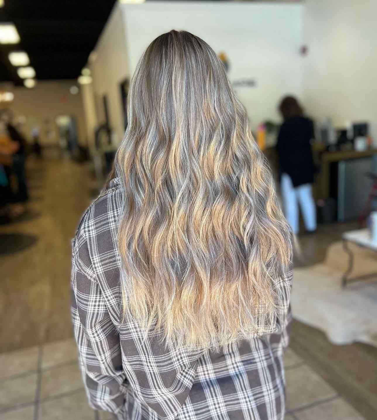 Person with long wavy hair wearing a plaid shirt, seen from behind in a salon interior.