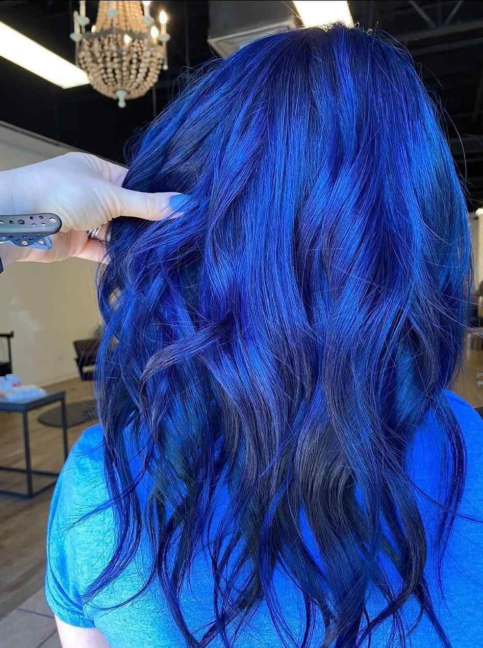 Back view of long wavy dark hair being held and styled in a salon chair under blue lighting