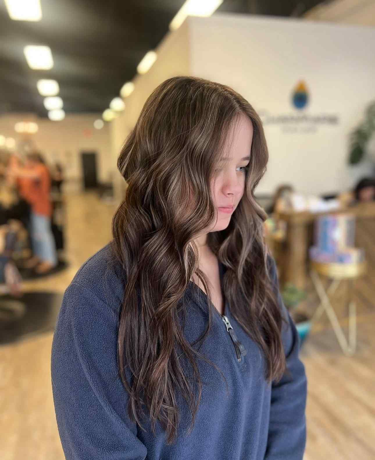 Woman with long wavy brown hair in a blue sweater, standing in a blurred indoor lobby.