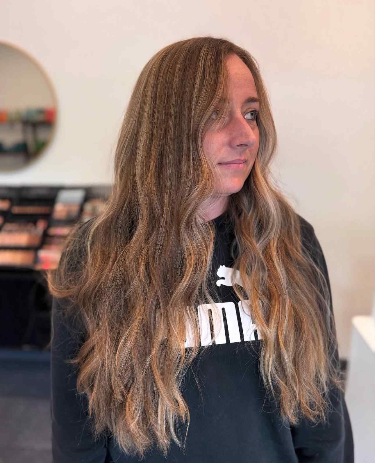 Person with long wavy hair wearing a black sweatshirt, standing indoors and looking to the side.