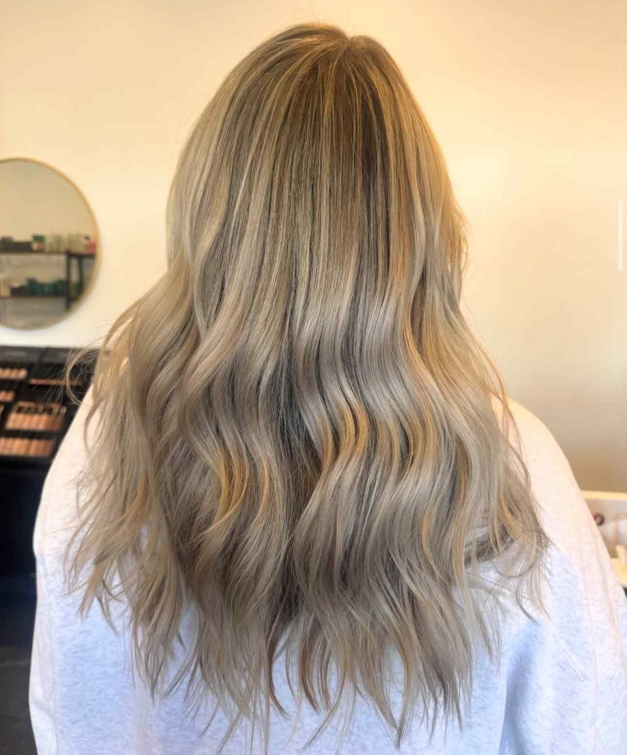 Back view of long wavy blonde hair with darker roots, falling over a light shirt indoors