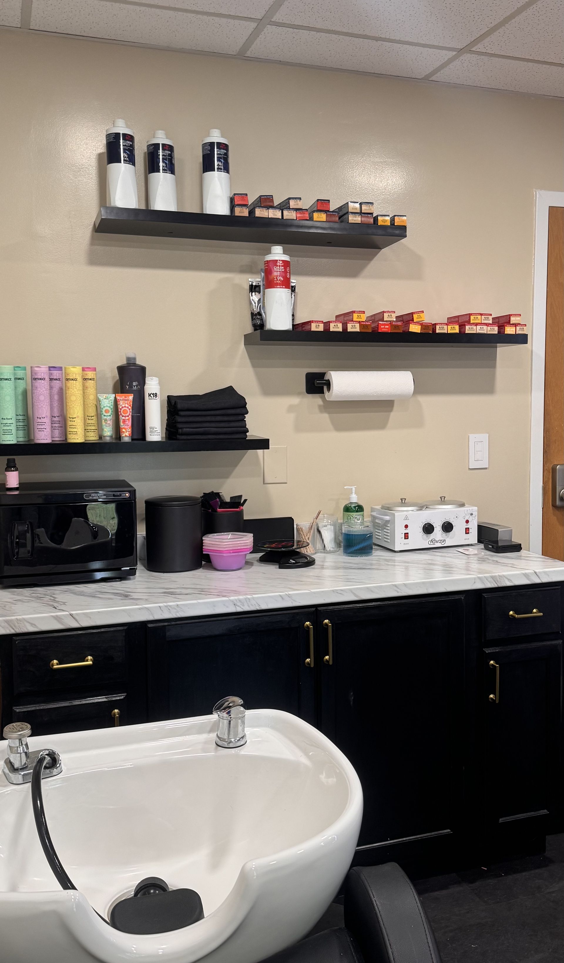 Salon sink and countertop with black cabinets, shelves of hair products, and a wall-mounted paper towel dispenser.