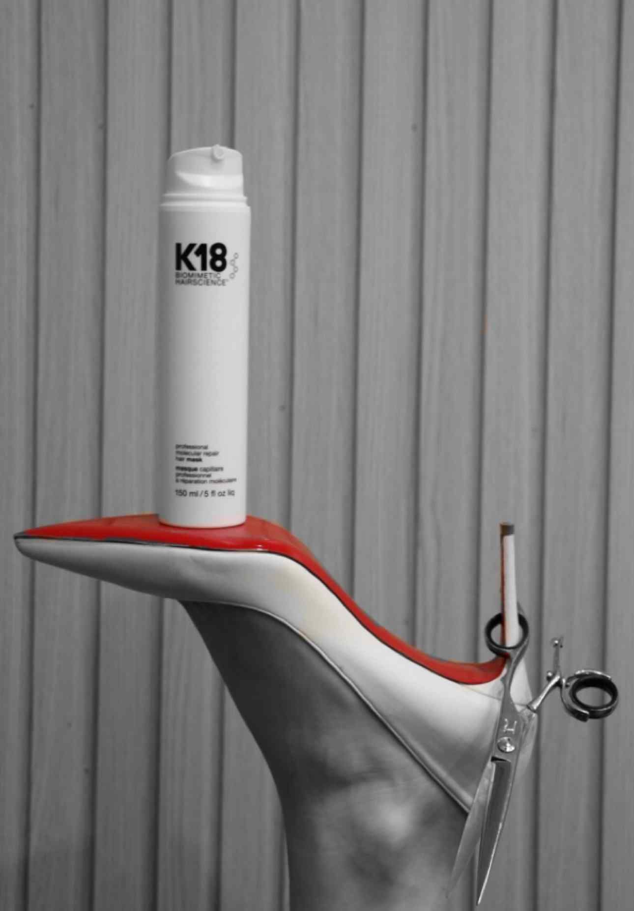 K18 hair product bottle balanced on a high heel shoe against a striped background