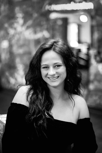 Smiling woman in an off-shoulder black top, posing outdoors with a blurred background.