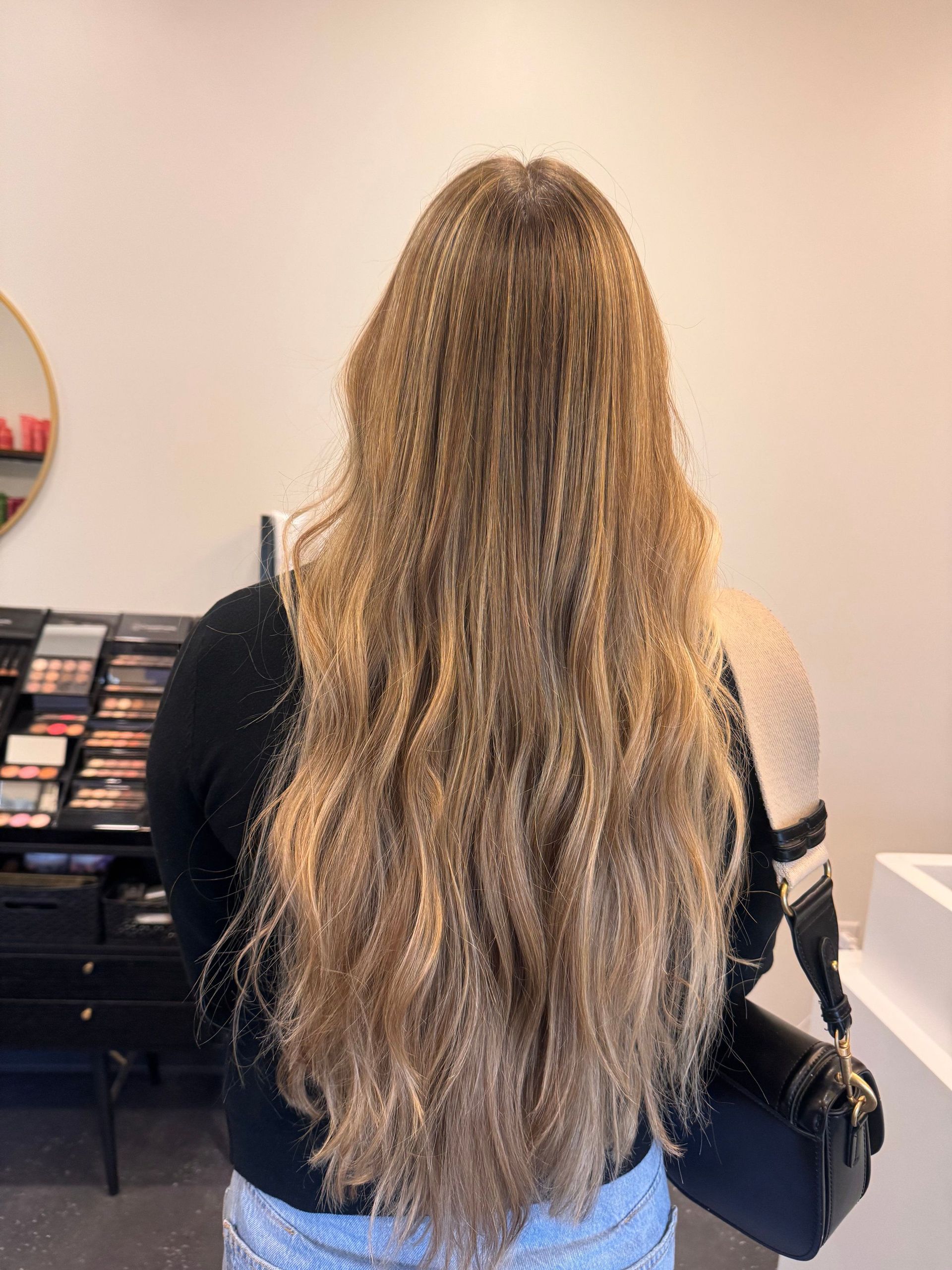 Person with long wavy blonde hair seen from behind, wearing a black top and carrying a black bag.