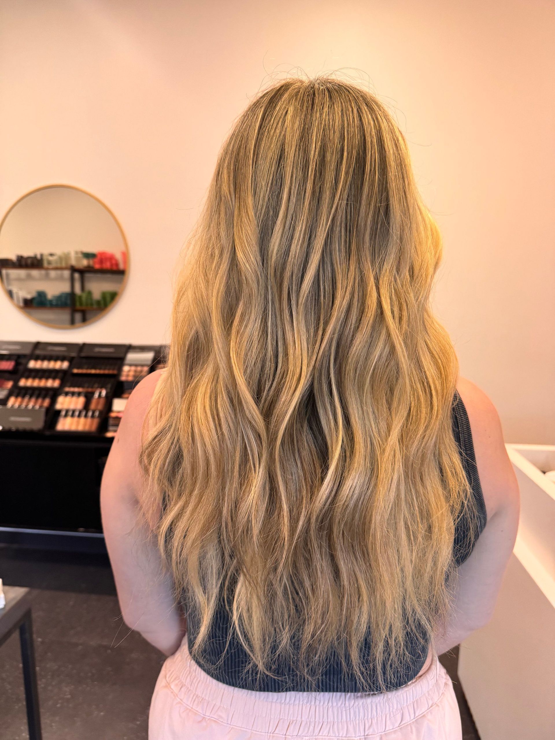 Person with long wavy blonde hair stands in a store, facing a makeup display.