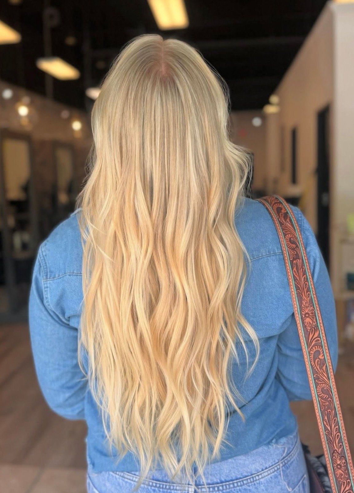 Person with long blonde hair in a blue shirt, seen from behind in a hallway with warm lights