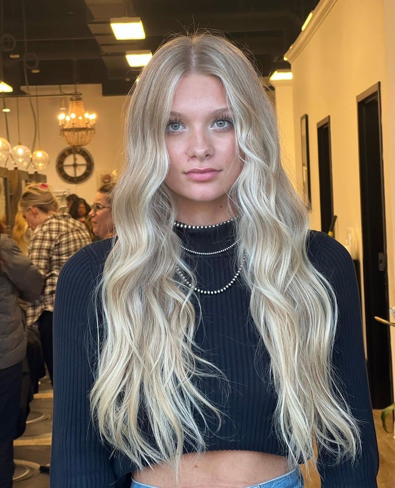 Young woman with long wavy blonde hair standing indoors in a black top and layered necklace.