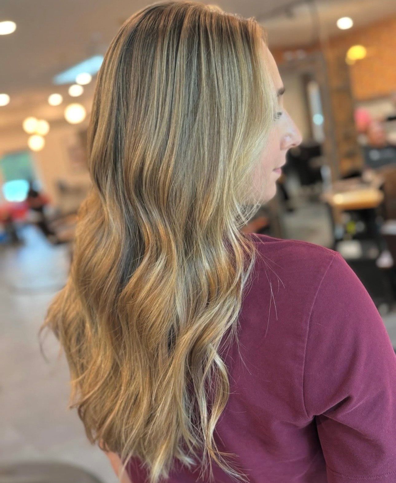 Woman with long wavy blonde hair in a maroon top, viewed from behind in a blurred indoor salon.