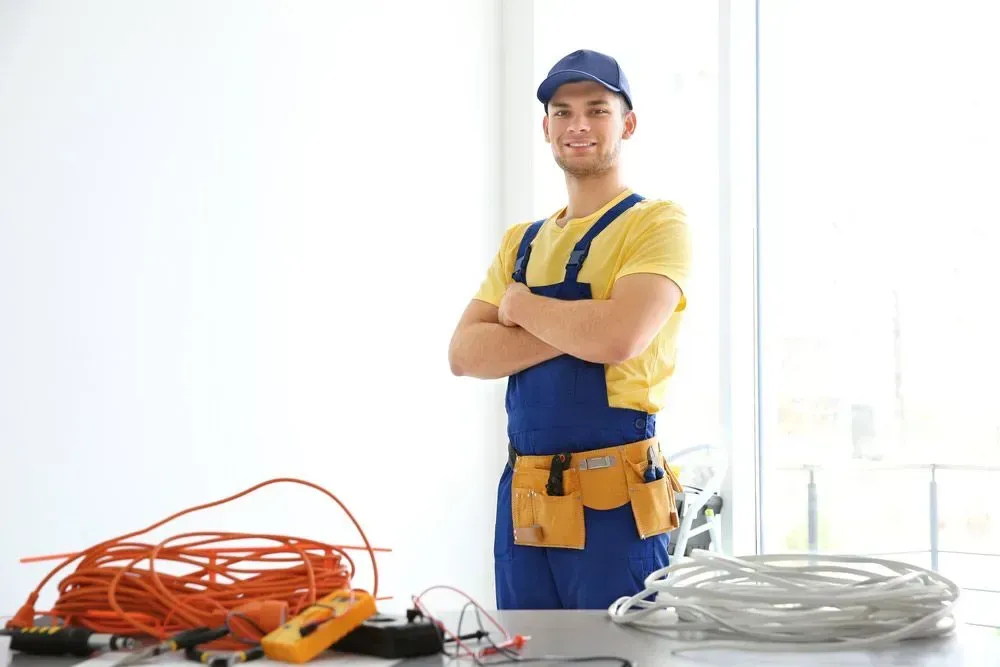 A Professional in a Yellow Shirt and Blue Overalls Stands Smiling — CH Electrics in Yeppoon, QLD