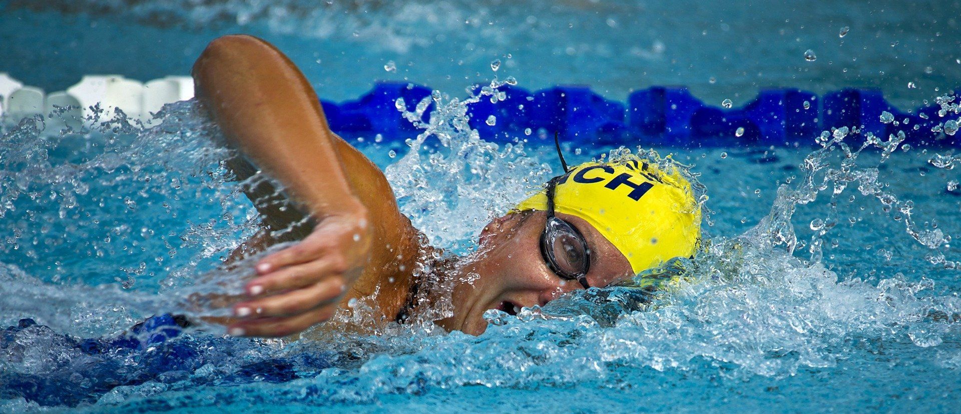 Swimmer in yellow cap doing freestyle in a blue pool, splashing water around the face