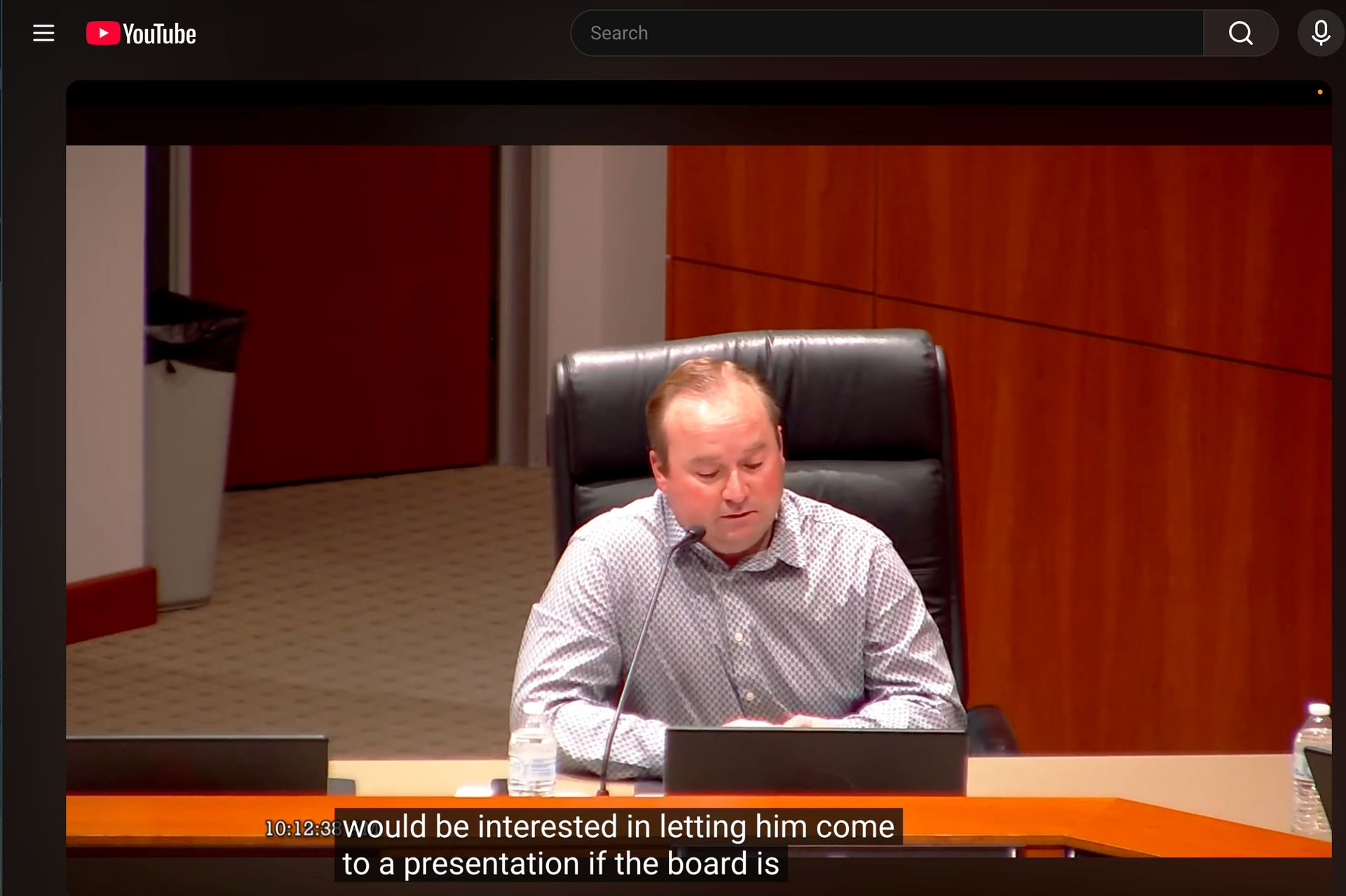 A man in a button-down shirt sits at a desk during a meeting with a YouTube overlay and subtitles at the bottom.