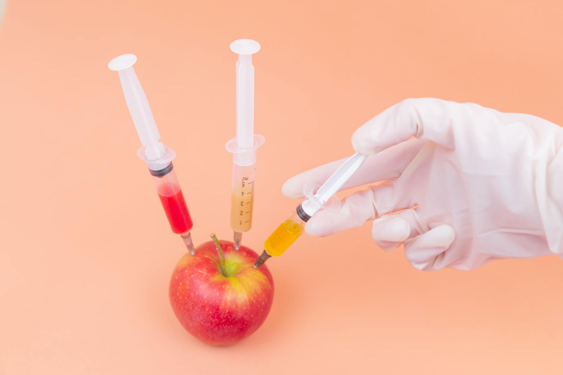 A gloved hand holds a syringe injecting a yellow liquid into a red apple, with two other syringes stuck into its top.