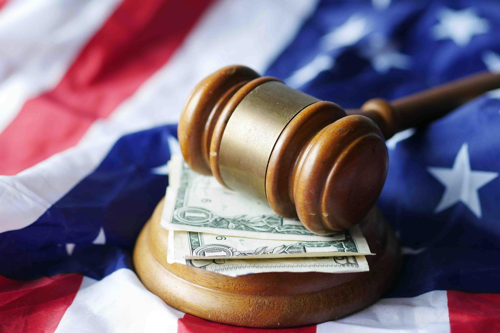 A wooden gavel resting on top of a small stack of US dollar bills, set against a background of an American flag.